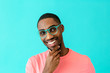 © Carlos David - Portrait of a happy young man with glasses smiling, thinking and looking to side with hand on chin, against blue studio background