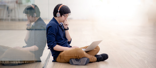 Asian man in casual clothing listening to music while sitting on the floor working with laptop computer in public building.  Urban lifestyle concept