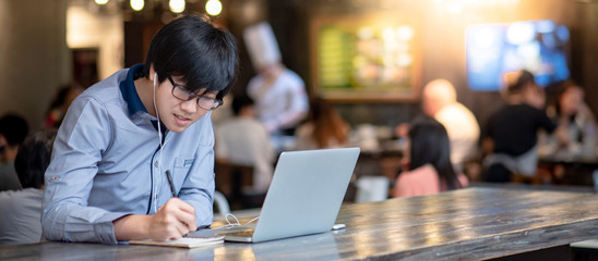 Asian businessman wearing glasses working with laptop computer and smartphone at restaurant. Male entrepreneur writing note while doing business talk. Technology for business management and strategy.