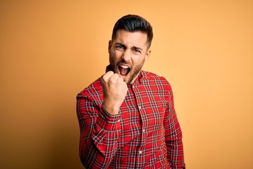 Canvas Print - Young handsome man wearing casual shirt standing over isolated yellow background angry and mad raising fist frustrated and furious while shouting with anger. Rage and aggressive concept.