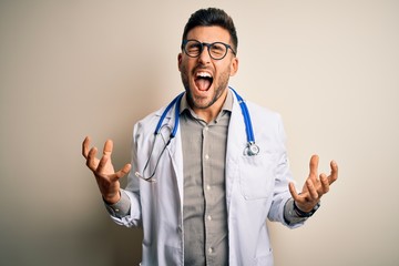 Canvas Print - Young doctor man wearing glasses, medical white robe and stethoscope over isolated background crazy and mad shouting and yelling with aggressive expression and arms raised. Frustration concept.