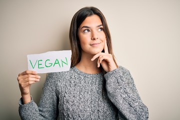 Canvas Print - Young beautiful girl holding paper with vegan message over isolated white background serious face thinking about question, very confused idea