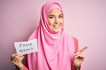 Sticker - Young beautiful girl wearing muslim hijab holding paper with fake news message very happy pointing with hand and finger to the side