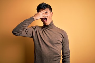 Poster - Young handsome man wearing casual turtleneck sweater over isolated yellow background peeking in shock covering face and eyes with hand, looking through fingers with embarrassed expression.