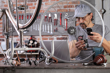 Wall Mural - man repair the vintage bicycle with mobile phone at the computer search spare parts on the internet in garage workshop on the workbench with tools, diy concept