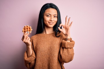 Young beautiful chinese woman holding waffle standing over isolated pink background doing ok sign with fingers, excellent symbol