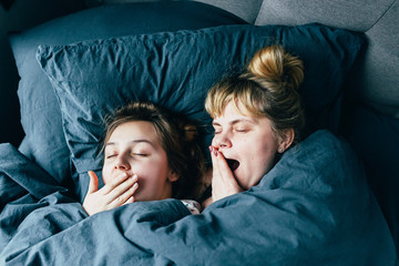 Portrait of light hair long hair mother and daughter on pillow under the duvet together yawn in soft sleepy morning light on blue linen bed. Concept of happy family living, relaxation, comfort, fun.