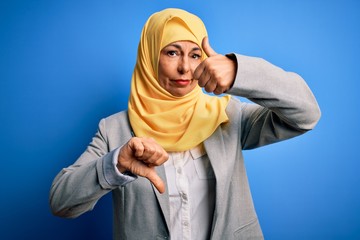 Wall Mural - Middle age brunette business woman wearing muslim traditional hijab over blue background Doing thumbs up and down, disagreement and agreement expression. Crazy conflict