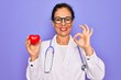 Middle age senior cardiologist doctor woman holding red heart over purple background doing ok sign with fingers, excellent symbol