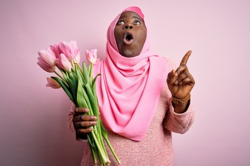 Canvas Print - Young african american plus size woman wearing muslim hijab holding bouquet of pink tulips amazed and surprised looking up and pointing with fingers and raised arms.