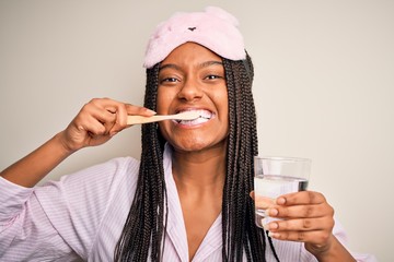 Canvas Print - Young beautiful african american woman wearing pajama and sleep mask brushing her teeth using tooth brush, oral paste and water glass, cleaning teeth and tongue as healthy health care morning routine