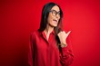 © Krakenimages.com - Young beautiful brunette woman wearing casual shirt and glasses over red background smiling with happy face looking and pointing to the side with thumb up.