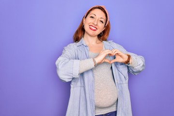 Wall Mural - Young beautiful redhead pregnant woman expecting baby over isolated purple background smiling in love doing heart symbol shape with hands. Romantic concept.