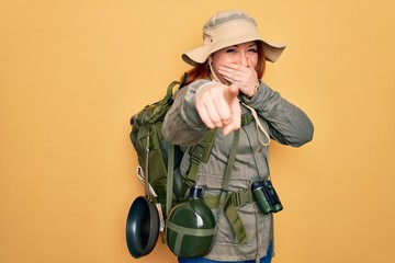 Sticker - Young redhead backpacker woman hiking wearing backpack and hat over yellow background laughing at you, pointing finger to the camera with hand over mouth, shame expression