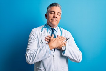 Poster - Middle age handsome grey-haired doctor man wearing coat and blue stethoscope smiling with hands on chest with closed eyes and grateful gesture on face. Health concept.