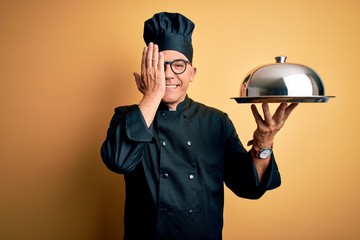 Poster - Middle age handsome grey-haired waiter man wearing cooker uniform and hat holding tray covering one eye with hand, confident smile on face and surprise emotion.