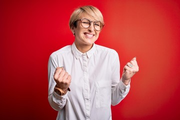 Wall Mural - Young blonde business woman with short hair wearing glasses over red background excited for success with arms raised and eyes closed celebrating victory smiling. Winner concept.