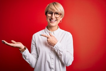 Wall Mural - Young blonde business woman with short hair wearing glasses over red background amazed and smiling to the camera while presenting with hand and pointing with finger.