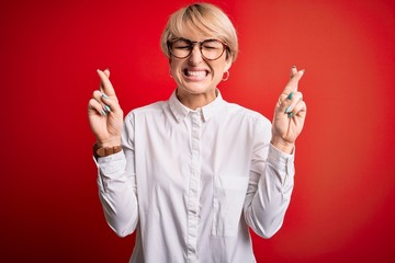 Poster - Young blonde business woman with short hair wearing glasses over red background gesturing finger crossed smiling with hope and eyes closed. Luck and superstitious concept.