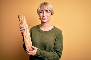 Sticker - Young blonde woman with short hair holding fresh bread baguette over yellow background with a confident expression on smart face thinking serious