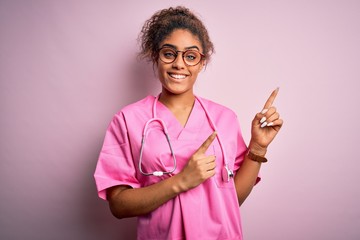 Poster - African american nurse girl wearing medical uniform and stethoscope over pink background smiling and looking at the camera pointing with two hands and fingers to the side.