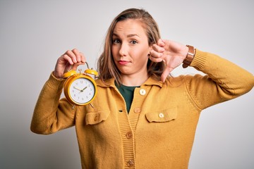 Poster - Young beautiful blonde woman holding alarm clock standing over isolated white background with angry face, negative sign showing dislike with thumbs down, rejection concept