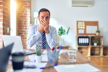 Wall Mural - Middle age handsome businessman wearing tie sitting using laptop at the office shocked covering mouth with hands for mistake. Secret concept.