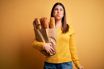 Canvas Print - Young beautiful woman holding a bag of fresh healthy bread over yellow background puffing cheeks with funny face. Mouth inflated with air, crazy expression.