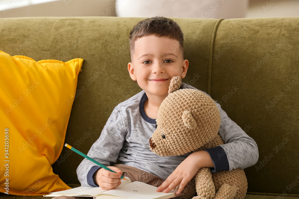 Cute little boy with teddy bear and notebook sitting on sofa at home