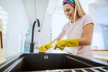 Worthy smiling dedicated housewife with rubber gloves on spraying and cleaning with sponge kitchen sink. Domestic kitchen interior.