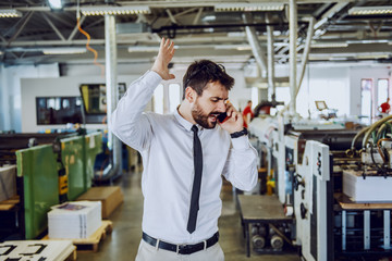 Nervous caucasian bearded graphic engineer talking on the phone and shouting. Printing shop interior.