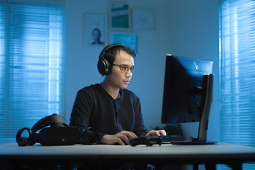 Casual Asian young man wearing black t-shirt  and headphone working from home with his computer and vr glasses at night with the warm blue light from the windows