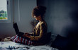 © Mlle Sonyah - Side view of a black woman working at home during Coronavirus quarantine. Conference call in the bedroom wearing pajamas and jacket. Dark cinematic photo.