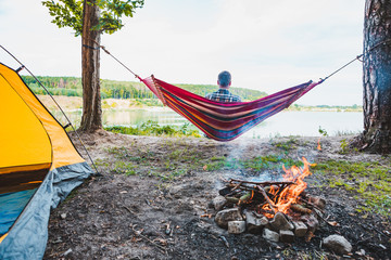 Wall Mural - man laying on hammock at lake beach near camp fire