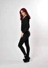 Wall Mural - Portrait of a pretty girl with red hair wearing black jeans, boots and a blouse.  full length standing pose in side profile,  on a studio background.