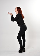 Sticker - Portrait of a pretty girl with red hair wearing black jeans, boots and a blouse.  full length standing pose in side profile,  on a studio background.