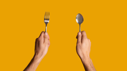 Unrecognizable millennial guy holding spoon and fork on orange background, close up. Panorama