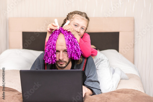 Telecommuting in quarantine. A fun family event. A little girl prevents dad from working at a computer. A child puts a bright wig on the head of a bald man.