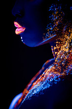 African Woman With Body Art Glowing In Ultraviolet Light. Portrait Of Beautiful Woman Painted In Fluorescent Powder. Isolated Dark Space