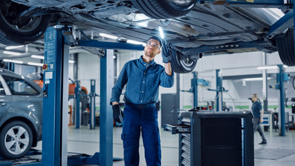 Wall Mural - Handsome Professional Car Mechanic is Working Under a Vehicle on a Lift in Service. Repairman is Using a LED lamp and Takes a Ratchet. Specialist is Wearing Safety Glasses. Modern Workshop.