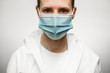 © fesenko - Close-up face of woman in medical mask and white protective suit which looks at camera.