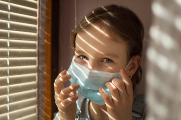 Little girl is sitting on the windowsill and trying to put protective disposable mask on her face during self-isolation at home because of Coronavirus Covid-19 on blinds background