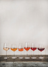 Various Shades Of Rose Wine In Stemmed Glasses Placed In Line From Light To Dark Colour, White Wall Background Behind, Copy Space. Wine Bar, Wine Shop, Wine Tasting Concept
