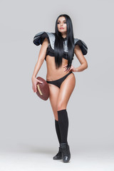 Wall Mural - Full length portrait of sexy brunette female american football player in uniform posing with a ball isolated on grey background
