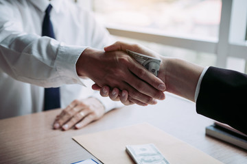 Two corporate businessmen shaking hands while one man places money on document in office room with corruption concept.