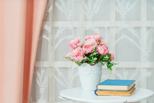Table With Flowers And Books On The Background Of A Window With Lace Tulle. Pink Flowers In Pots In The Interior. Artificial Pink Fake Flowers In A White Vase.