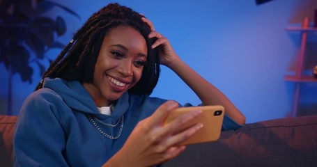Millennial attractive girl watching video while holding her phone in horizontal landscape mode. Young woman with dreadlocks and nose ring using smartphone and smiling at home