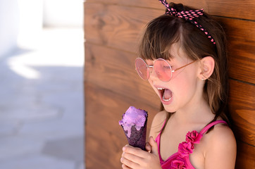 Closeup of pretty little girl eating ice cream outdoors on sunny day. Cute girl in pink swimsuit licking purple ice-cream in waffle cone. Summer, happy childhood concept. Copy space for your text