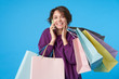 © timtimphoto - Joyful young attractive brown-eyed curly brunette lady smiling while showing her pleasant emotions during phone talk, standing over blue background with paper bags in raised hands