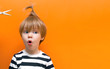 © Natal.is - little european blond child boy with long hair and crest opened his mouth in surprise and looks towards the text. concept of a hairdresser and hairstyles for children. Isolated on orange background.